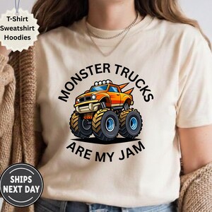 May include: Beige t-shirt with a cartoon monster truck graphic. The orange and yellow truck has large tires and the text "MONSTER TRUCKS ARE MY JAM" in black. The shirt also has a "T-Shirt Sweatshirt Hoodies" label.