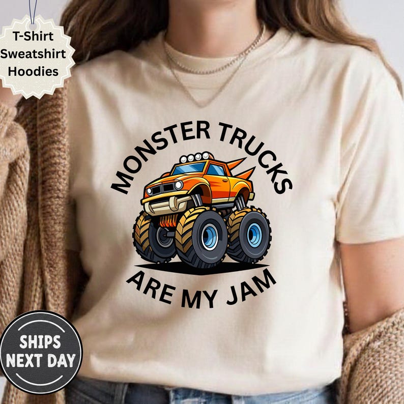 May include: Beige t-shirt with a cartoon monster truck graphic. The orange and yellow truck has large tires and the text "MONSTER TRUCKS ARE MY JAM" in black. The shirt also has a "T-Shirt Sweatshirt Hoodies" label.