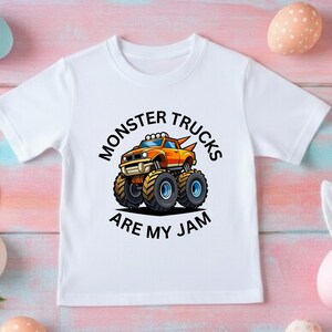May include: White t-shirt featuring a cartoon monster truck in orange and yellow with black tires. The text "MONSTER TRUCKS ARE MY JAM" is printed in black around the truck.