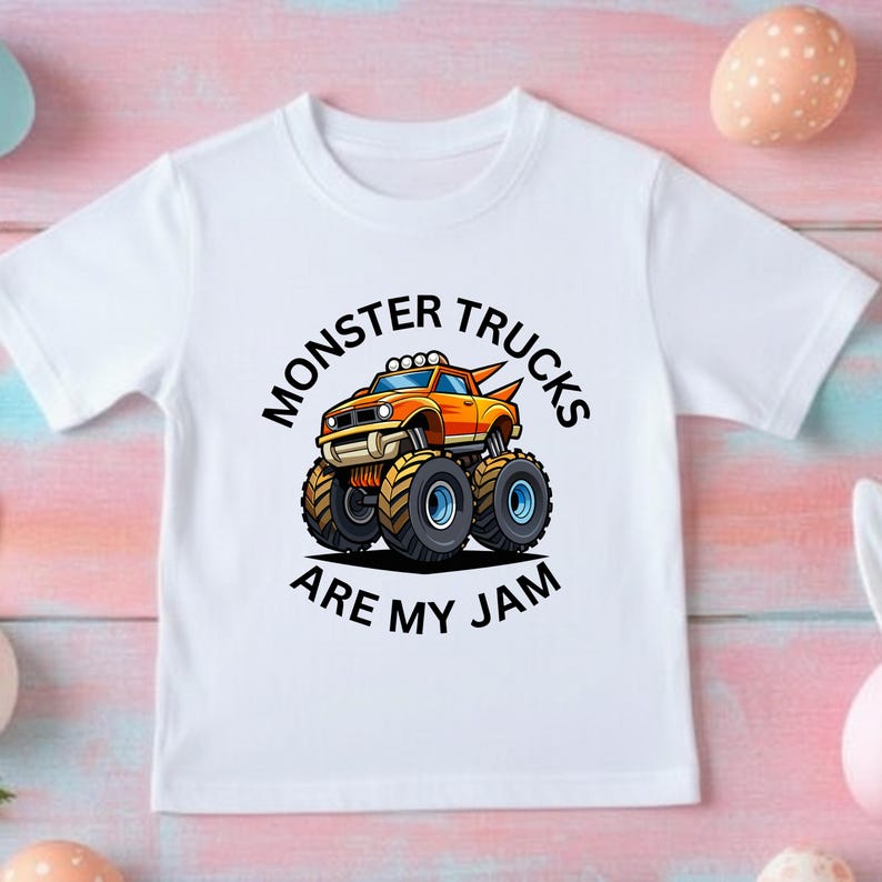 May include: White t-shirt featuring a cartoon monster truck in orange and yellow with black tires. The text "MONSTER TRUCKS ARE MY JAM" is printed in black around the truck.