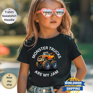 May include: A black t-shirt featuring a cartoon monster truck design in orange, yellow, and blue. The text "MONSTER TRUCKS ARE MY JAM" is printed above and below the truck graphic. The shirt is worn by a child.