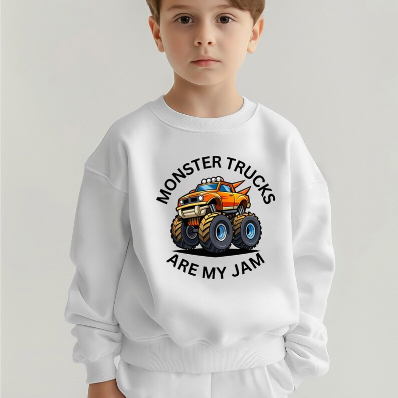 May include: White sweatshirt featuring a colorful cartoon monster truck design. The orange, blue, and yellow truck has large tires and the text "MONSTER TRUCKS ARE MY JAM" in a circular layout.