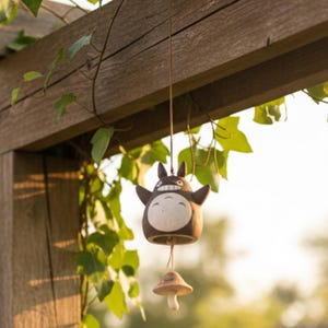 Puede incluir: Campanilla de viento de cerámica marrón con un personaje de Totoro sonriente con los brazos extendidos y un badajo en forma de hongo. La campanilla cuelga de una viga de madera, rodeada de hojas verdes, creando una decoración exterior caprichosa.