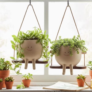 Op de afbeelding: Twee beige keramische hangplantenbakken met lachende gezichten, elk met groene planten. De plantenbakken zijn met bruin touw aan een metalen haak opgehangen. De plantenbakken hebben poten en staan op een kleine houten plank.