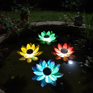 Puede incluir: Cuatro luces flotantes solares con forma de flor de loto en un estanque. Las flores son amarillas, verdes, rojas y azules, cada una con un panel solar negro en el centro. Las luces están encendidas, creando una colorida exhibición en el agua.