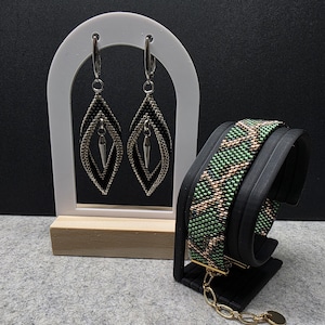 Op de afbeelding: Een paar ruitvormige kralenoorbellen met zwarte en zilveren kraaltjes en een zilveren puntvormige bedel, naast een brede kralenarmband met een groen, zwart en goudkleurig geometrisch patroon en een goudkleurige kettingverlenger.