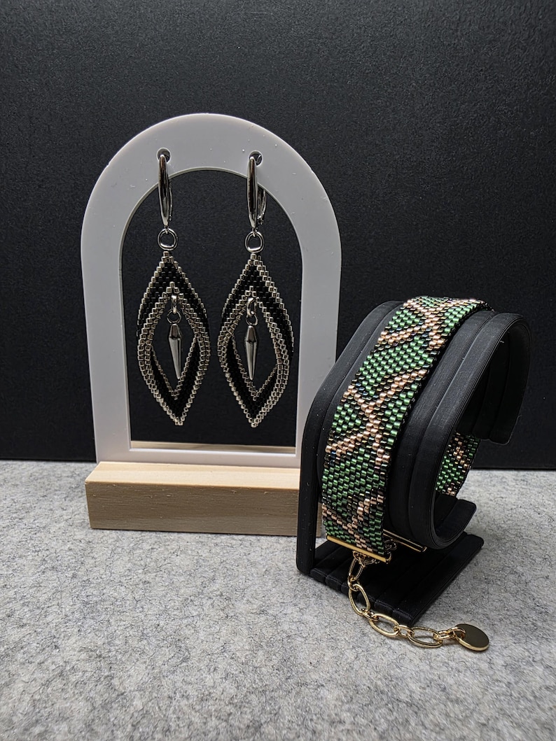 Op de afbeelding: Een paar ruitvormige kralenoorbellen met zwarte en zilveren kraaltjes en een zilveren puntvormige bedel, naast een brede kralenarmband met een groen, zwart en goudkleurig geometrisch patroon en een goudkleurige kettingverlenger.