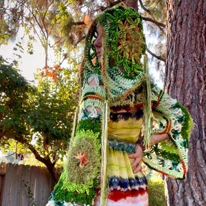 May include: A colorful, handmade crochet outfit. The ensemble includes a green and white crochet top with bell sleeves and a matching head covering. The dress is a tie-dye pattern of yellow, red, blue, and white. The outfit is worn outdoors.