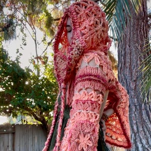 May include: A handmade, open-shoulder crochet hooded shawl in shades of peach, coral, and burgundy. The shawl features a floral pattern on the sleeves and hood, with long braided ties. The garment is worn over a green dress.