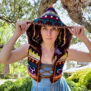 May include: A woman wearing a colorful crocheted vest with a hood. The vest has a brown base with multi-colored stripes and a lace-up front. The hood is pulled up over her head and has a similar pattern to the vest.