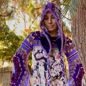 May include: A handmade crochet hooded jacket in shades of purple and lavender. The jacket features intricate openwork and textured details on the sleeves and hood. The model is wearing a floral print dress. The overall aesthetic is bohemian and artistic.