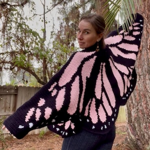 Puede incluir: Un suéter de ala de mariposa de ganchillo negro y rosa. El suéter presenta alas grandes y detalladas con patrones rosas y negros. El diseño se asemeja a una mariposa monarca. El suéter se usa al aire libre.