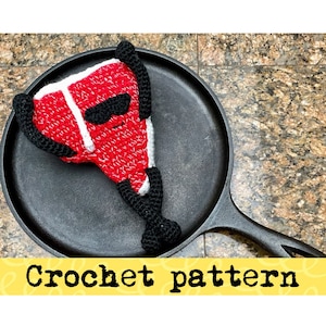 Peut inclure: Un steak en crochet rouge et blanc avec des yeux noirs et une poignée en crochet noir. Le steak est posé sur une poêle en fonte noire.