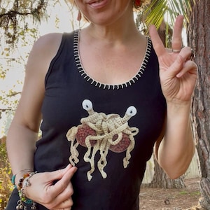 May include: Black vest top with a white stitched neckline and a beige and red graphic of the Flying Spaghetti Monster. The top is sleeveless and made of a soft material.
