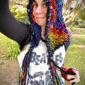 May include: A colorful crocheted hooded scarf with long fringe tassels. The scarf is made with black, red, orange, yellow, green, blue, and purple yarn. The scarf is worn over a white t-shirt with a Beatles graphic.