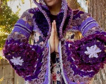 May include: A handmade crochet hooded jacket in shades of purple, lavender, and gray. The jacket features a floral design on the sleeves and hood, with a fringed trim. The garment is worn over a floral print dress.