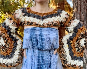 May include: A woman wearing a crocheted top with long bell sleeves in shades of brown, white, and gray. The top has a wide, open neckline and a loose fit. The woman is standing in front of a tree with a blue and white tie-dye skirt.