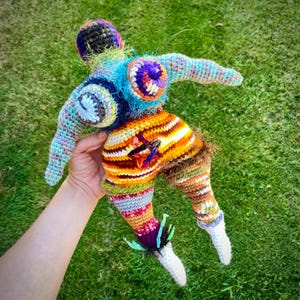 May include: A handmade, colorful crochet doll with a whimsical design. The doll features a multi-colored body, arms, and legs, with a textured, yarn-like appearance. The doll's head is topped with a dark, textured cap, and the legs have white feet. The doll is held against a green background.