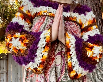May include: A woman wearing a crocheted sweater with purple, orange, and white yarn. The sweater has a bell sleeve design and a crocheted trim. The woman is standing in front of a tree.