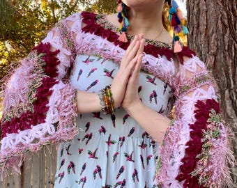 May include: A woman wearing a light blue dress with a pink and burgundy crochet top. The dress has a hummingbird print. She is wearing colorful tassel earrings and bracelets.