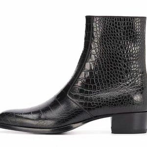 Peut inclure: Une bottine noire avec un motif texturé en forme de peau de crocodile. La botte a un bout pointu, une fermeture éclair latérale et un petit talon bloc. La botte est faite d'un matériau brillant semblable à du cuir.