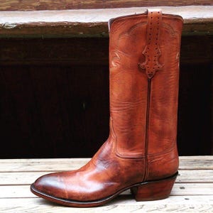 May include: A tall, brown leather cowboy boot with a pointed toe and a stacked heel. The boot features decorative stitching and a pull strap at the top. The leather has a rich, aged appearance, with variations in color and texture.