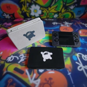 Op de afbeelding: Een assortiment gamingaccessoires, waaronder een handheld console en twee hoesjes. De hoesjes zijn rechthoekig, één lichtbeige en de andere zwart, elk met een cartoon ninja logo. De handheld console is donkergrijs met een scherm en knoppen.