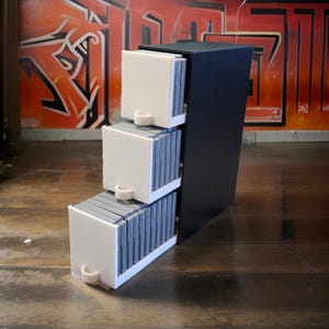 Può includere: Un'unità di stoccaggio nera a gradini con tre cassetti bianchi. Ogni cassetto contiene una pila di cartucce di gioco grigie. L'unità è posizionata contro un muro coperto di graffiti.