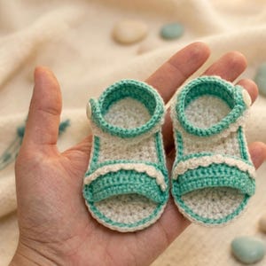 May include: A pair of handmade crochet baby sandals. The sandals are white and teal, with a button closure. The sandals have a scalloped edge and are designed for a baby. The sandals are made of soft yarn.