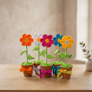 Könnte beinhalten: Sechs handgefertigte Häkelblumen in kleinen Terrakottatöpfen. Jede Blume hat eine andere Farbe: orange, weiß, pink, blau und gelb. Grüne Stiele und Blätter tragen die Blumen. Die Töpfe sind mit bunten Bändern verziert.