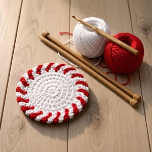 May include: A round, white and red crocheted coaster with a spiral pattern, accompanied by two wooden knitting needles, a white yarn ball, and a red yarn ball, with a wooden crochet hook resting across the top of the white yarn ball.