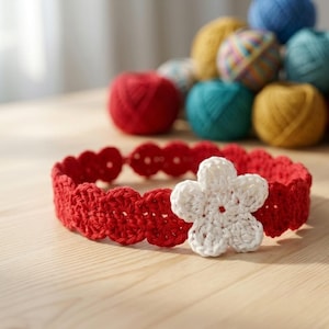 Puede incluir: Una diadema roja de ganchillo con un adorno de flor blanca. La diadema tiene un borde festoneado y está hecha de hilo texturizado. En el fondo, hay ovillos de lana de colores, lo que sugiere un proyecto de manualidades.