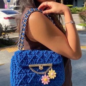 May include: A bright blue crochet handbag with a gold chain strap and clasp. The bag has a decorative chain with two flower charms in pink and yellow. The bag's design includes a diamond pattern.