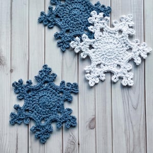 May include: Three crocheted snowflake ornaments, two in a muted blue yarn and one in a bright white yarn. Each snowflake features a circular center with intricate, branching points radiating outward in a classic six-sided geometric pattern.