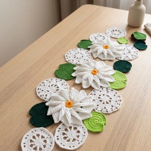 May include: A handmade crochet table runner featuring white daisy-like flowers with yellow centres, green lily pads, and white doilies. The runner is arranged on a light wooden surface, creating a decorative and floral theme.