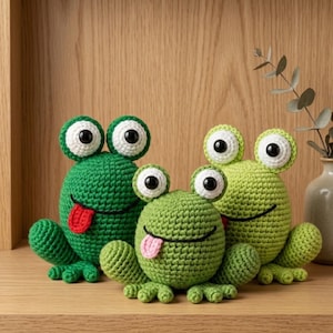 May include: Three crocheted frog figurines in varying shades of green. The frogs have large, white-rimmed eyes with black pupils and protruding tongues in red or pink. They are arranged on a wooden surface, suggesting a decorative or handmade item.
