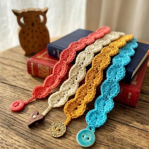 May include: Four crochet bookmarks in coral, cream, yellow, and turquoise. Each bookmark has a wavy design and a button or wooden heart accent. The bookmarks are displayed on a wooden surface with books in the background.