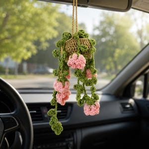 May include: A crocheted hanging plant ornament featuring a tan basket, green trailing vines, and pink flower blossoms. The decorative item is suspended by a tan cord and is designed to hang from a vehicle rearview mirror for interior display.