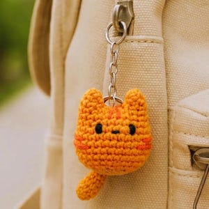 Op de afbeelding: Een gehaakte oranje katten sleutelhanger met zwarte ogen en een kleine staart. De kat heeft oranje strepen op zijn hoofd en gezicht. De sleutelhanger is bevestigd aan een zilveren ketting en een rits.