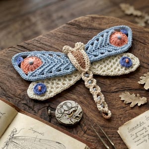 May include: A handcrafted crochet dragonfly in shades of blue, cream, and peach, resting on a wooden surface. The dragonfly features intricate openwork wings and a detailed body. Decorative elements include a vintage watch mechanism and botanical illustrations.
