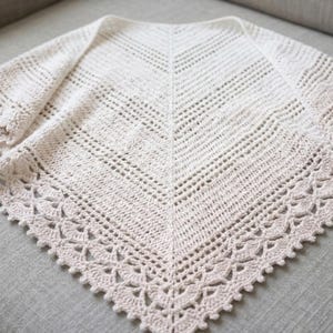 May include: A cream-colored, crocheted shawl with a triangular shape. The shawl features a detailed openwork design throughout, with a decorative scalloped edge. The texture and pattern are clearly visible, highlighting the handmade quality of the item.