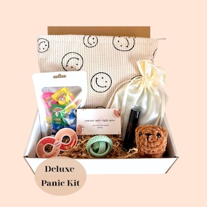 Könnte beinhalten: Eine weiße Box mit Artikeln zur Selbstpflege. Enthält eine cremefarbene Tasche mit Smiley-Gesichtern, eine Tüte mit bunten Süßigkeiten, ein kleines braunes Stofftier, eine Rollerflasche und eine Karte mit der Aufschrift "you are safe right now". Die Box ist mit "Deluxe Panic Kit" beschriftet.