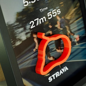 Könnte beinhalten: Gerahmter Druck mit einem verschwommenen Bild von Läufern und dem Text "5:5" und "27m 55s". Eine leuchtend orangefarbene, stilisierte Form ist vorne angebracht. Das Wort "STRAVA" ist unten aufgedruckt.