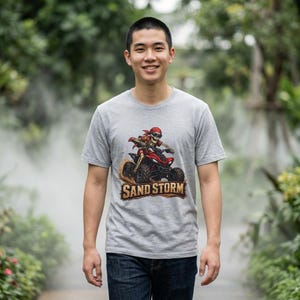May include: A gray t-shirt featuring a graphic of a skeleton riding a red ATV through sand. The text "SAND STORM" is displayed below the ATV. The person in the image is wearing blue jeans.