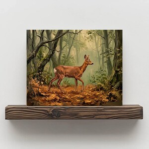 May include: A framed painting depicts a deer walking through a forest. The artwork features a brown deer with a light brown coat, set against a backdrop of trees and fallen leaves. The color palette includes shades of brown, green, and yellow.