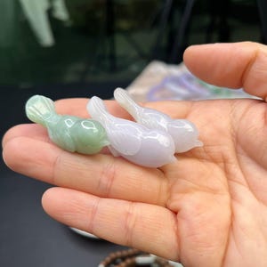 Puede incluir: Tres pequeñas figuras de pájaros talladas en jade. Una es de color verde claro, y las otras dos son de un lavanda pálido. Los pájaros son lisos y pulidos, con un diseño simple y elegante. Estas figuras podrían usarse como objetos decorativos o de colección.