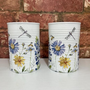 May include: Two cylindrical metal containers painted white, each featuring a hand-painted botanical design. The artwork displays blue and yellow flowers, green stems, small bees, and dragonflies scattered across the ribbed exterior surface.