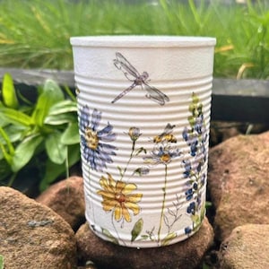 May include: A white, upcycled tin can planter decorated with a floral and dragonfly design. The design features blue, yellow, and white flowers, along with a dragonfly and bees. The can is sitting on a bed of brown rocks.