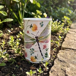 May include: A decorated tin can featuring a dragonfly and floral pattern. The dragonfly has a black body and wings in shades of purple, green, and pink. The flowers are yellow and pink, set against a white background.