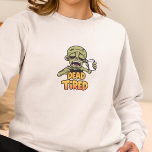 K&ouml;nnte beinhalten: Hellgraues Sweatshirt mit einem Cartoon-Zombie, der eine Kaffeetasse h&auml;lt. Der Zombie hat gr&uuml;ne Haut und einen m&uuml;den Gesichtsausdruck. Die Worte "DEAD TIRED" sind darunter in Orange und Gelb gedruckt.